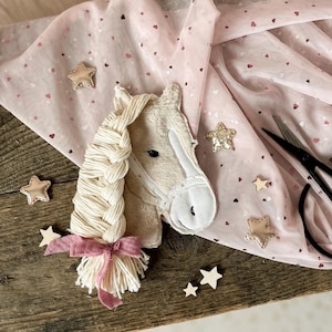 Könnte beinhalten: Ein cremefarbener Plüschpferdekopf mit geflochtener Mähne und rosa Schleife. Goldene Sterne und ein rosa Stoff mit Herzmustern sind verstreut. Das Pferd hat einen weißen Zaum.