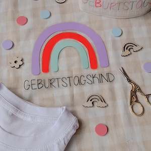 Könnte beinhalten: Ein weißes T-Shirt mit dem Text "Geburtstagskind" und einem Regenbogen-Design. Außerdem sind eine Krone, eine Blume und eine Schere auf dem Bild zu sehen.