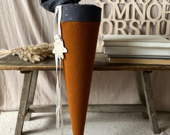 May include: A brown and orange fabric cone with a white ribbon and a wooden rocket-shaped ornament. The cone is topped with a dark blue fabric with gold stars. The cone is sitting on a wooden table with a stack of books behind it.