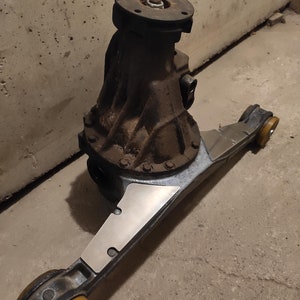 Op de afbeelding: Een roestige metalen achterasdifferentiaal met twee gele rubberen bevestigingen. Het differentieel is bevestigd aan een metalen frame met twee zilveren metalen platen.