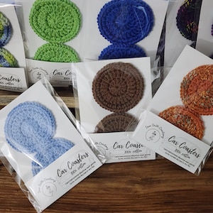May include: A collection of handmade car coasters in various colors, including green, blue, brown, and orange. Each coaster is made of 100% cotton and packaged in clear plastic. The packaging includes the text "Car Coasters" and care instructions.