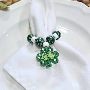 May include: A white napkin ring with green and white beads and a green shamrock charm with the word "Lucky" on it. The napkin ring is on a white plate.