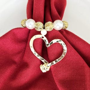 May include: A gold-colored heart-shaped napkin ring with a textured finish, hanging from a beaded ring of white and gold beads. The napkin ring is set against a folded red napkin.