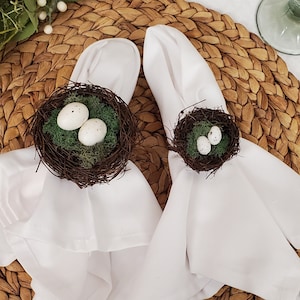 May include: Two white cloth napkins with brown bird's nest napkin rings decorated with white eggs and green moss. The napkins are on a woven brown placemat.