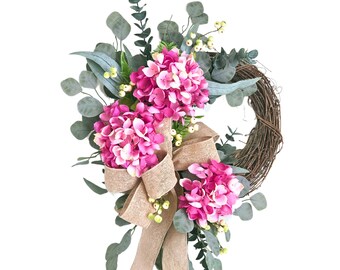 May include: A pink and green floral wreath with a burlap bow. The wreath is made of grapevine and features pink hydrangeas and eucalyptus leaves.