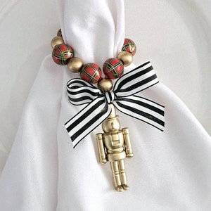 May include: A white napkin ring with a gold nutcracker charm and red and green plaid beads. The napkin ring is on a white plate with a red charger.