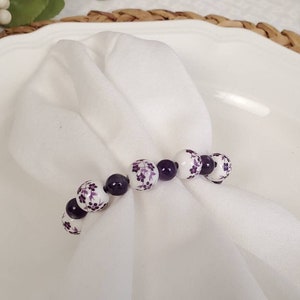 May include: White napkin ring with purple and white floral beads. The beads are strung together to form a circle.