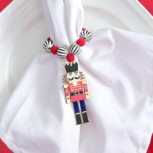 May include: A white napkin with a red and white beaded nutcracker ring, placed on a red and white plate with a green garland around the edge.
