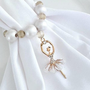 May include: A white napkin held by a beaded napkin ring. The ring features large, white faux pearls and gold-colored beads. A gold-colored ballerina charm with clear rhinestones and white accents hangs from the ring.