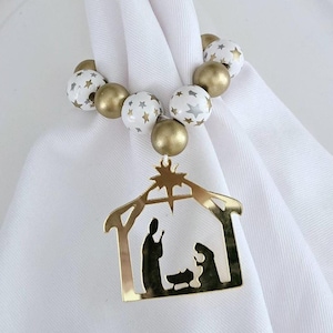 May include: White napkin with a gold nativity scene ornament and a gold and white beaded napkin ring.