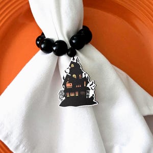 May include: A white cloth napkin held by a black beaded napkin ring. A Halloween-themed house-shaped charm with bats and a tree hangs from the ring. The background includes an orange plate.
