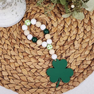 May include: A green shamrock charm hangs from a beaded garland. The garland is made of white, beige, and green beads with a checkered pattern. The garland is on a woven brown surface.