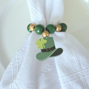 May include: A white napkin is held by a beaded napkin ring. The ring features green and gold beads. A green leprechaun hat with a shamrock charm hangs from the ring. The hat has a black brim and a brown belt.