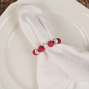 May include: Red and white baseball themed napkin ring. The ring is made of wooden beads painted red with white baseball stitching.