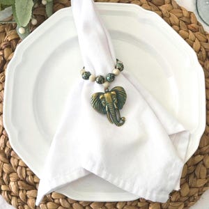 May include: A white cloth napkin held by a beaded napkin ring with a bronze elephant pendant. The napkin is on a white plate with a scalloped edge, set on a woven placemat.