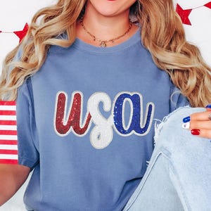 以下が含まれることがあります： 赤、白、青のキラキラした「USA」のグラフィックが施された青いTシャツです。