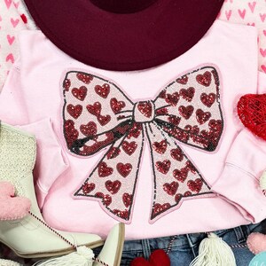 May include: A light pink sweatshirt with a large sequined bow design filled with red hearts. A burgundy hat, white boots, and heart-shaped decorations are arranged around the sweatshirt. The Sweet Tee Doll logo is visible.