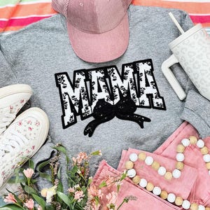 May include: A gray sweatshirt with the word "MAMA" in black and white floral lettering. The sweatshirt is paired with a pink corduroy hat and a wooden bead necklace.