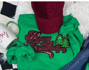 Puede incluir: Una sudadera verde con la frase "Merry Christmas" en lentejuelas rojas y dos árboles de Navidad verdes con lentejuelas. También se ve una gorra de béisbol de pana granate y una taza blanca con la palabra "Joy".