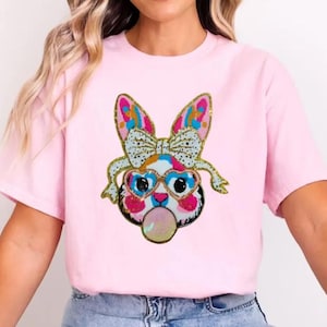 May include: A light pink t-shirt with a cartoon bunny face design. The bunny has a bow, glasses, and a bubble gum bubble. The design includes gold glitter accents. The shirt is made of a soft material.