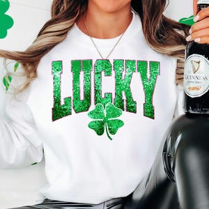 May include: White sweatshirt with the word "LUCKY" in green glitter lettering and a green shamrock. The person is holding a Guinness beer bottle. The background has green shamrock decorations.