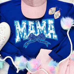 Op de afbeelding: Een koninklijk blauw sweatshirt met een witte en blauwe bloemenprint "MAMA" graphic met een blauwe glitter strik. Het sweatshirt is gecombineerd met een roze corduroy pet en een wit touw met veelkleurige pompoenen.
