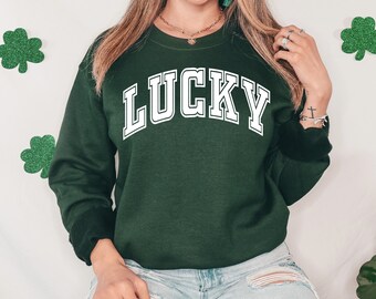 May include: A dark green sweatshirt with the word "LUCKY" in white, block letters. The sweatshirt has a crew neck and long sleeves. The image also features green glitter shamrock decorations.