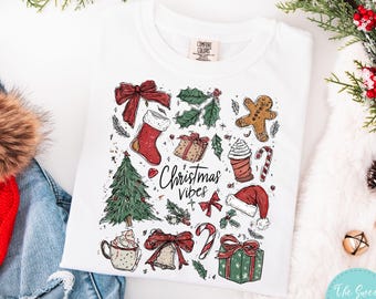 May include: White t-shirt featuring a festive Christmas design. The graphic includes a Christmas tree, stocking, gifts, gingerbread man, and the words "Christmas Vibes". The shirt is set against a backdrop of seasonal decorations.