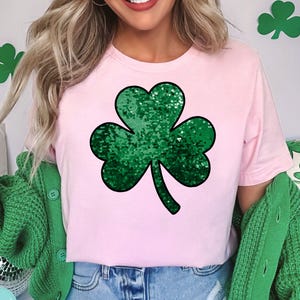 Op de afbeelding: Een lichtroze t-shirt met een groot, groen pailletten klaverblad. Het klaverblad is het centrale ontwerp. De persoon draagt een groen vest. De achtergrond heeft klaverbladdecoraties.