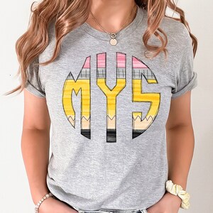 Könnte beinhalten: Graues T-Shirt mit einem gelben, rosa und grauen Bleistiftdesign, das "MYS" buchstabiert.