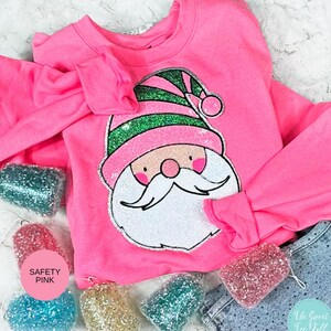 May include: A bright pink sweatshirt with a Santa Claus graphic. The Santa has a white beard, pink cheeks, and a pink and green hat. The sleeves are flared. Several glittery ornaments in various colors are scattered around the shirt. The text "SAFETY PINK" is visible.