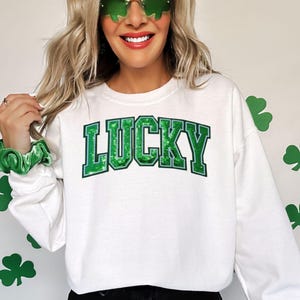 Könnte beinhalten: Weißes Sweatshirt mit dem Wort "LUCKY" in großen grünen Blockbuchstaben. Das Wort ist in einem dunkleren Grün umrandet. Das Sweatshirt wird mit schwarzen Shorts kombiniert. Grüne Kleeblattdekorationen befinden sich an der Wand.
