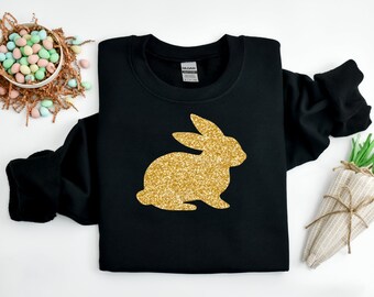 Può includere: Felpa nera con un design a coniglio glitterato dorato. Il coniglio è una silhouette. L'immagine include anche decorazioni pasquali, come un cestino di uova dai colori pastello e una carota decorativa.