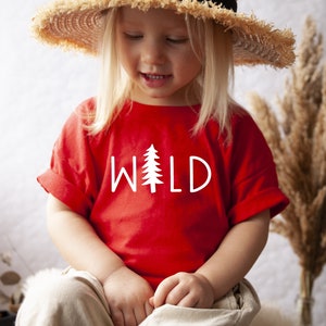 Może przedstawiać: Małe dziecko w czerwonej koszulce z napisem "WILD" i grafiką białej sosny. Dziecko nosi słomkowy kapelusz i ma blond włosy.