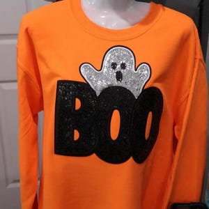 Może przedstawiać: Jasnopomarańczowa bluza z cekinowym napisem "BOO" i błyszczącą grafiką ducha. Top z długim rękawem jest prezentowany na manekinie, idealny na Halloween.