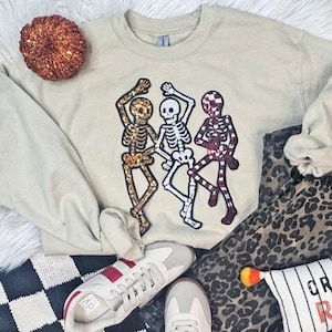 May include: A beige sweatshirt with three dancing skeleton figures in gold, white, and burgundy. The image includes a decorative pumpkin, trainers, leopard print trousers, and a striped pillow with the word "CREEP" on it.