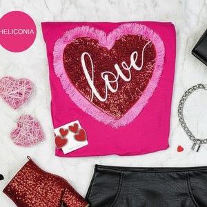Könnte beinhalten: Eine Draufsicht mit Kleidung und Accessoires zum Valentinstag. Ein pinkes Shirt mit einem paillettenbesetzten Herz und dem Wort "love", ein schwarzer Rock, rote Glitzerstiefel, eine schwarze Handtasche und eine silberne Halskette.