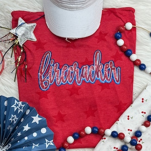 May include: Red t-shirt with a sparkly blue and red "firecracker" design. The shirt is decorated with white stars and is surrounded by red, white, and blue decorations.