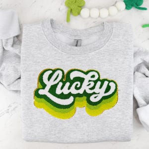 Könnte beinhalten: Ein hellgraues Sweatshirt mit dem Wort "Lucky" in einem geschichteten Retro-Design. Der Text ist weiß mit grünen und gelben Akzenten und einer goldenen Umrandung. Das Sweatshirt ist auf einer weißen Oberfläche gefaltet.