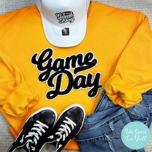 Może przedstawiać: Żółta bluza z czarnym nadrukiem "Game Day" i biała czapka bejsbolówka z napisem "Game Day". Bluza jest zestawiona z niebieskimi dżinsami i czarnymi butami sportowymi.