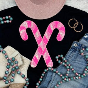 May include: Black t-shirt featuring a sequined pink candy cane design. The image also includes a pink hat, gold hoop earrings, blue jeans, and beaded necklaces. White boots are also visible.