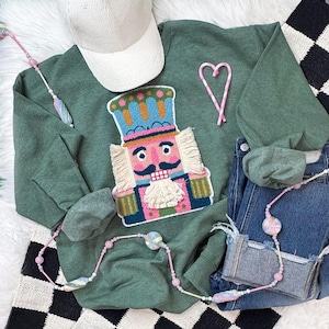 May include: Green sweatshirt with a colorful nutcracker applique, a white corduroy cap, and a pair of blue jeans. A pink and white candy cane heart and a beaded necklace are also visible. The items are arranged on a white fluffy surface.