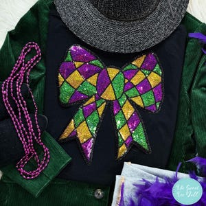 May include: Black long-sleeved top featuring a large sequined bow in green, gold, and purple. The image includes a green corduroy jacket, a grey hat, and a strand of pink beads. The Sweet Tee Yall logo is visible.