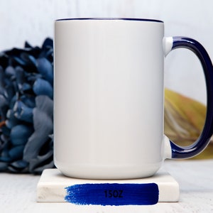 May include: White ceramic mug with a blue rim and handle. The mug is sitting on a white coaster with a blue stripe and the text "15oz" printed on it.