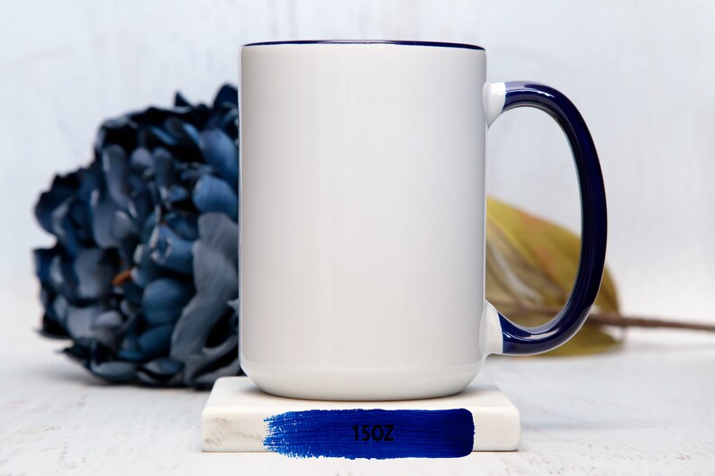May include: White ceramic mug with a blue rim and handle. The mug is sitting on a white coaster with a blue stripe and the text "15oz" printed on it.