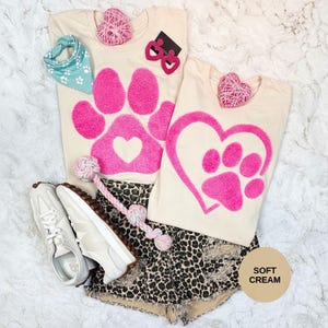 Op de afbeelding: Twee crèmekleurige t-shirts met roze pootafdrukontwerpen, gecombineerd met een luipaardprint short, witte sneakers en een hondenspeeltje. Accessoires: een turquoise bandana en roze hartvormige oorbellen.