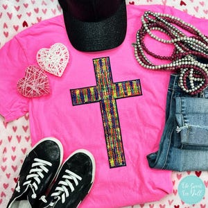 May include: A pink t-shirt with a colorful cross design, paired with black sneakers, a black cap, denim jeans, and beaded necklaces. Decorative white and pink heart-shaped ornaments are also present. The Sweet Tee Yall logo is visible.