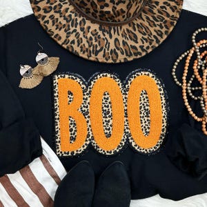 May include: Black sweatshirt featuring the word "BOO" in orange letters with a leopard print pattern. The image includes a leopard print hat, earrings, and a necklace. Black shoes are visible at the bottom.