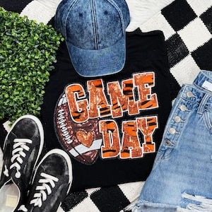 May include: A flat lay featuring a black t-shirt with "GAME DAY" text and a football graphic. The shirt is paired with denim shorts, a denim baseball cap, and black sneakers. The background includes a black and white checkered pattern.