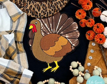 May include: Black t-shirt with a brown and gold sequined turkey applique. The shirt is surrounded by plaid fabric, a leopard print hat, and a string of brown and green beads.